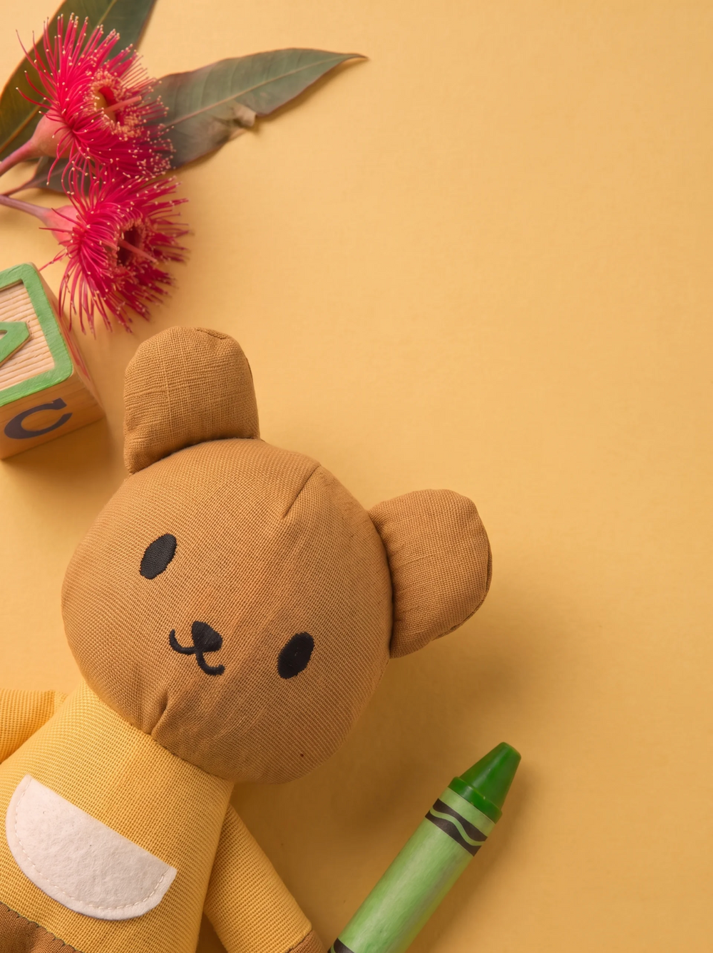 A plush kangaroo toy made from tan linen-like fabric for kindergarten children gifts, arranged on a warm beige background with a green crayon, pink flowers, and a wooden block nearby.