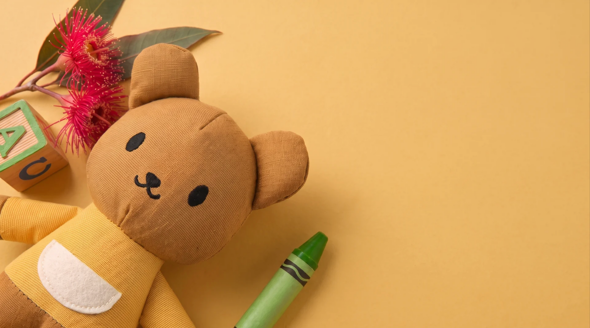 A linen kangaroo toy wearing a yellow shirt, styled on a solid yellow background with a green crayon, pink Australian eucalyptus flowers, and wooden preschool blocks.